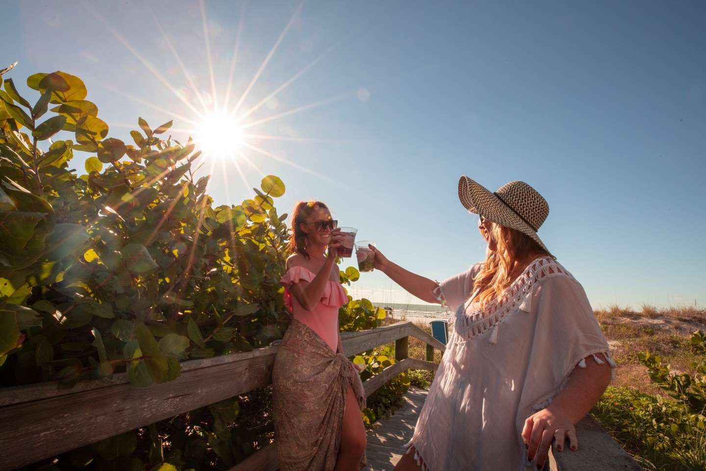 Two women on a small bridge to the beach tapping their glasses together as they walk past each other