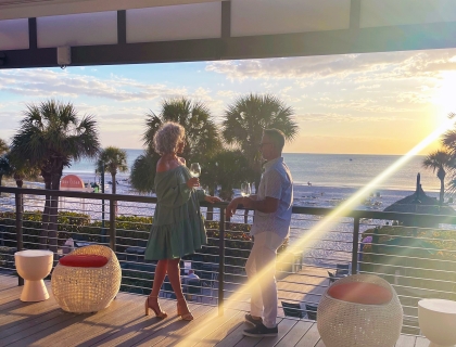 A couple stands on a balcony, gazing at the beach and ocean waves under a clear blue sky.
